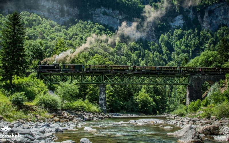 Ein Dampfzug überquert eine grüne Metallbrücke über einen felsigen Fluss, der von dichten grünen Wäldern und steilen Klippen umgeben ist.