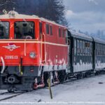 Eine rote und cremefarbene Diesellokomotive zieht mehrere grüne Personenwagen über verschneite Gleise durch eine winterliche Landschaft mit Hügeln und Bäumen im Hintergrund.