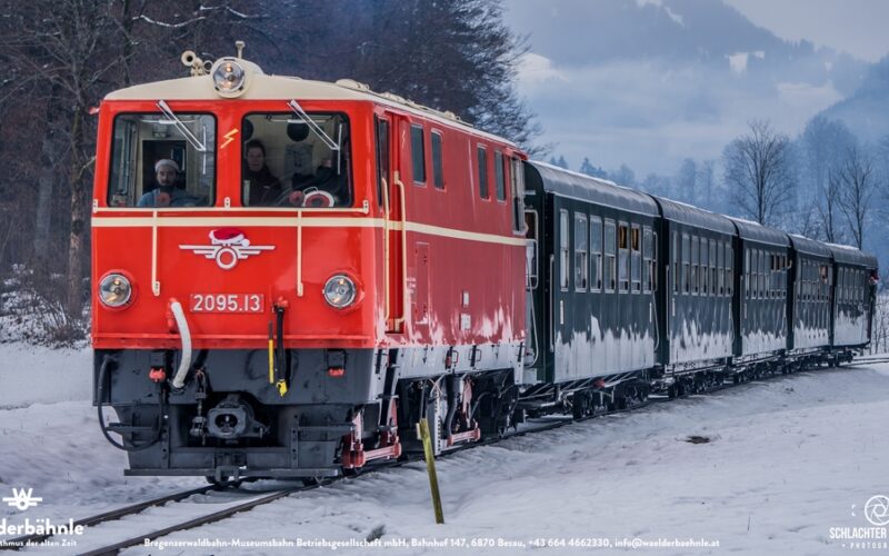 Eine rote und cremefarbene Diesellokomotive zieht mehrere grüne Personenwagen über verschneite Gleise durch eine winterliche Landschaft mit Hügeln und Bäumen im Hintergrund.