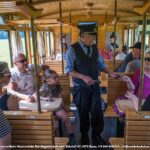 Die Fahrgäste sitzen in einem hölzernen Zugwaggon, während ein Schaffner die Fahrkarten kontrolliert; Menschen unterschiedlichen Alters sitzen zusammen und unterhalten sich.