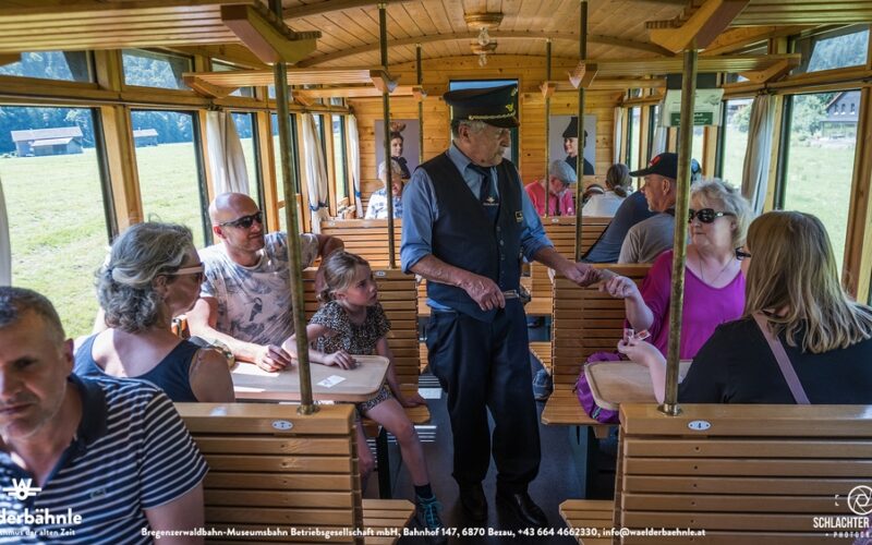 Die Fahrgäste sitzen in einem hölzernen Zugwaggon, während ein Schaffner die Fahrkarten kontrolliert; Menschen unterschiedlichen Alters sitzen zusammen und unterhalten sich.
