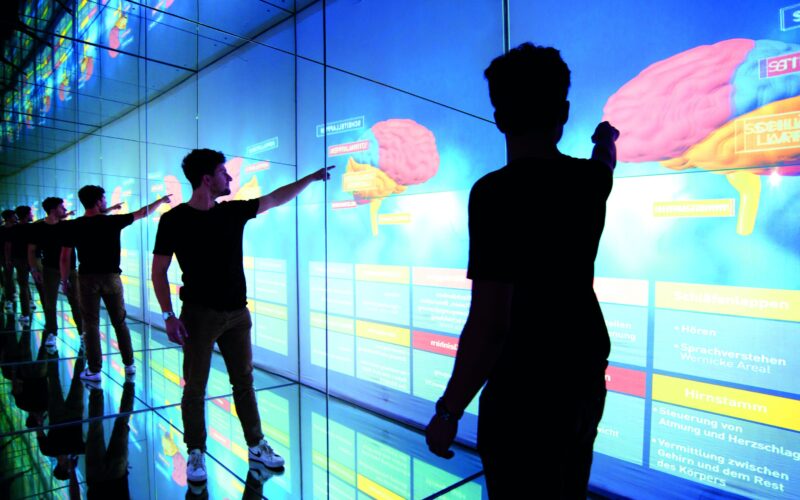 Eine Person in einem dunklen T-Shirt zeigt auf ein großes digitales Display, auf dem ein Gehirn abgebildet ist, während sich die Szene an den verspiegelten Wänden und auf dem Boden widerspiegelt.