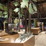 Museumsexponat mit Holzbalken, didaktischen Schautafeln, Tiermodellen, Blattschmuck und Besuchern, die die Exponate in einem großen Raum betrachten.