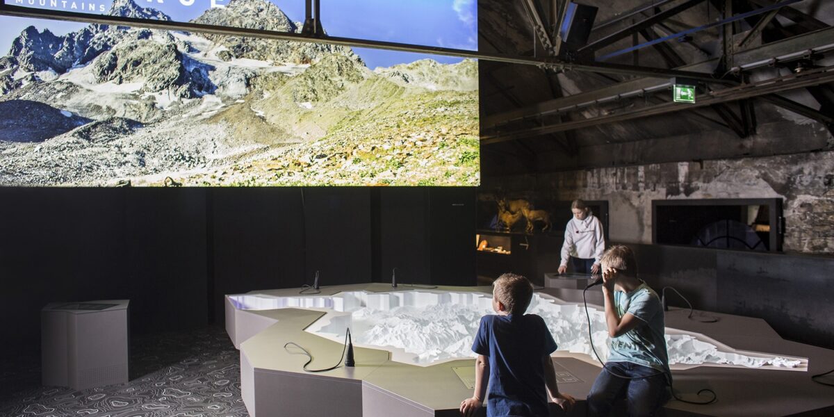 Zwei Kinder betrachten ein großes 3D-Geländemodell unter einem Display, das eine Gebirgslandschaft mit dem Wort "GEBIRGE" in einem modernen Ausstellungsraum zeigt.