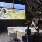Zwei Kinder betrachten ein großes 3D-Geländemodell unter einem Display, das eine Gebirgslandschaft mit dem Wort "GEBIRGE" in einem modernen Ausstellungsraum zeigt.
