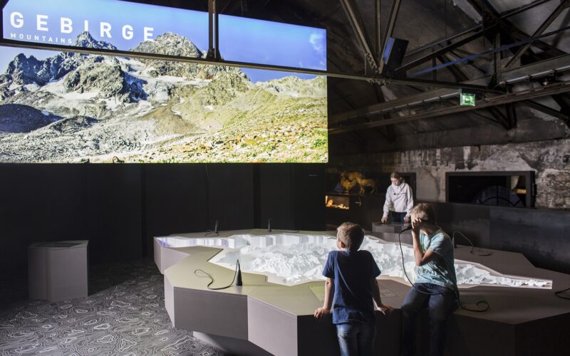 Zwei Kinder betrachten ein großes 3D-Geländemodell unter einem Display, das eine Gebirgslandschaft mit dem Wort "GEBIRGE" in einem modernen Ausstellungsraum zeigt.
