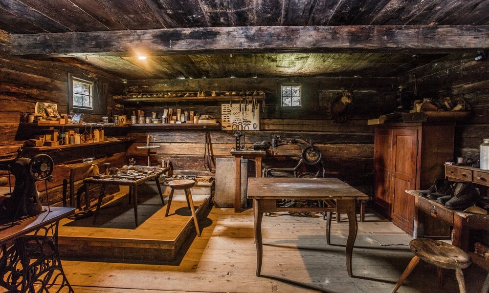 Das Innere einer alten Holzwerkstatt mit alten Werkzeugen, Tischen, Regalen, einer Nähmaschine und kleinen Fenstern, die Tageslicht hereinlassen.