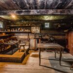 Das Innere einer alten Holzwerkstatt mit alten Werkzeugen, Tischen, Regalen, einer Nähmaschine und kleinen Fenstern, die Tageslicht hereinlassen.