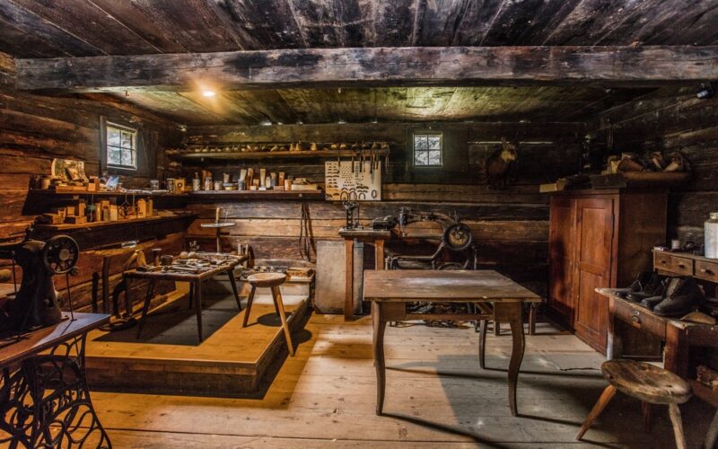 Das Innere einer alten Holzwerkstatt mit alten Werkzeugen, Tischen, Regalen, einer Nähmaschine und kleinen Fenstern, die Tageslicht hereinlassen.