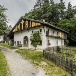 Ein rustikales weißes Haus mit einem Holzdach, gelbe Maiskolben, die unter dem Dachvorsprung trocknen, umgeben von Grün und einem kleinen Garten, entlang eines Kiesweges.