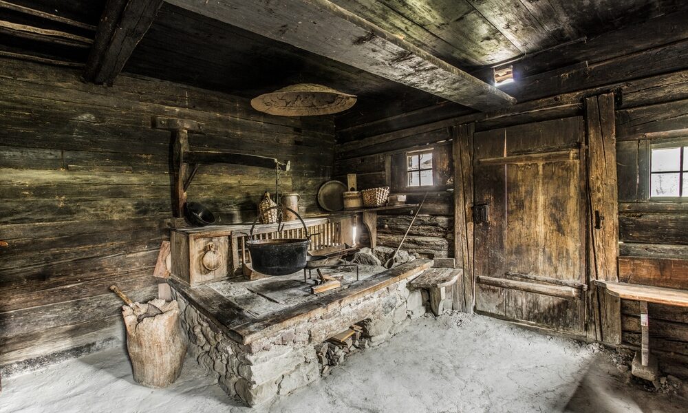 Interieur einer alten, rustikalen Holzhüttenküche mit Steinherd, hängenden Töpfen, Holzutensilien und geflochtenen Körben auf Regalen.