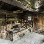 Interieur einer alten, rustikalen Holzhüttenküche mit Steinherd, hängenden Töpfen, Holzutensilien und geflochtenen Körben auf Regalen.