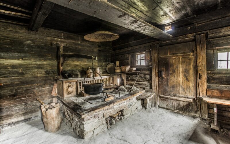 Interieur einer alten, rustikalen Holzhüttenküche mit Steinherd, hängenden Töpfen, Holzutensilien und geflochtenen Körben auf Regalen.