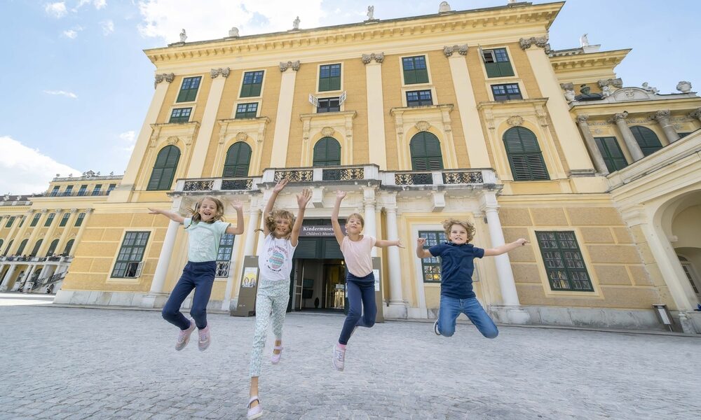 Vier Kinder springen vor einem großen, gelben historischen Gebäude mit vielen Fenstern und Säulen in die Luft.