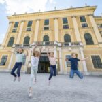 Vier Kinder springen vor einem großen, gelben historischen Gebäude mit vielen Fenstern und Säulen in die Luft.