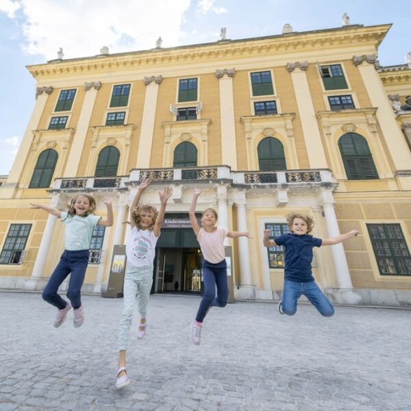 Vier Kinder springen vor einem großen, gelben historischen Gebäude mit vielen Fenstern und Säulen in die Luft.