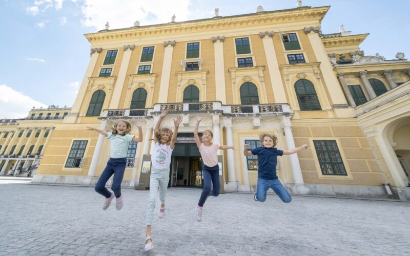Vier Kinder springen vor einem großen, gelben historischen Gebäude mit vielen Fenstern und Säulen in die Luft.