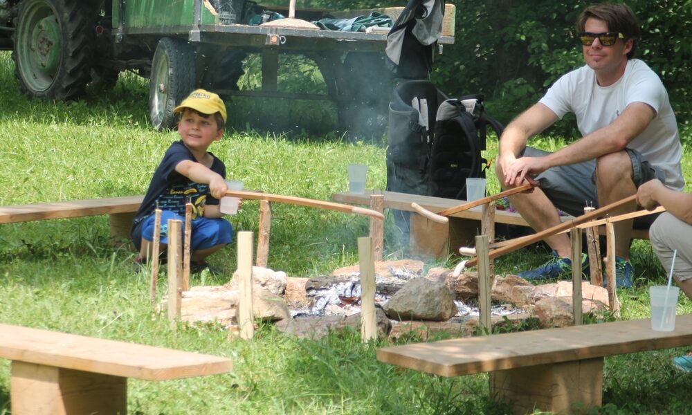 Ein Junge und ein Erwachsener rösten Essen auf Stöcken über einem Lagerfeuer, umgeben von Holzbänken, mit einem Traktor und einem Wagen im Hintergrund.