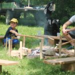 Ein Junge und ein Erwachsener rösten Essen auf Stöcken über einem Lagerfeuer, umgeben von Holzbänken, mit einem Traktor und einem Wagen im Hintergrund.