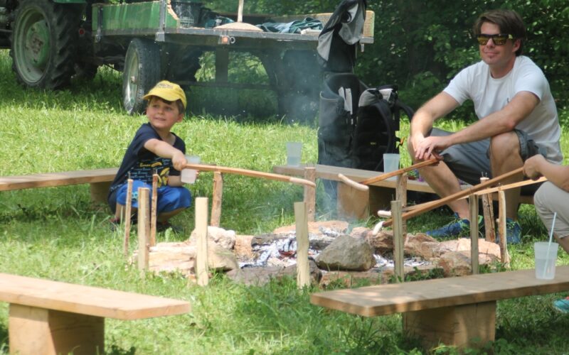 Ein Junge und ein Erwachsener rösten Essen auf Stöcken über einem Lagerfeuer, umgeben von Holzbänken, mit einem Traktor und einem Wagen im Hintergrund.