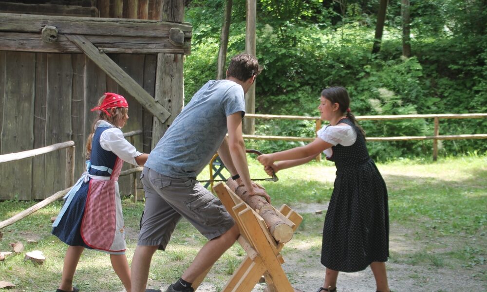 Zwei Mädchen in traditioneller Kleidung und ein Mann sägen mit einer Zweiersäge einen Baumstamm im Freien in der Nähe einer Holzkonstruktion und eines Zauns.