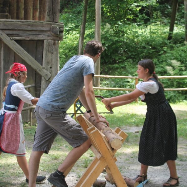 Zwei Mädchen in traditioneller Kleidung und ein Mann sägen mit einer Zweiersäge einen Baumstamm im Freien in der Nähe einer Holzkonstruktion und eines Zauns.