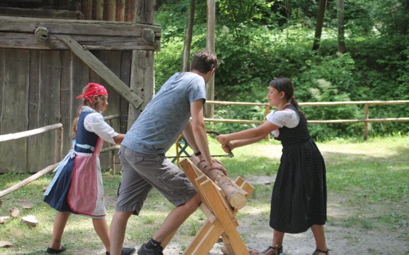 Zwei Mädchen in traditioneller Kleidung und ein Mann sägen mit einer Zweiersäge einen Baumstamm im Freien in der Nähe einer Holzkonstruktion und eines Zauns.