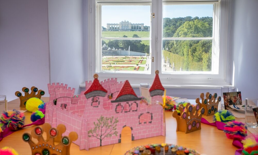 Auf einem Tisch stehen eine rosa Pappburg, verzierte Kronen und Bastelmaterial. Durch das Fenster hat man einen Blick auf einen angelegten Garten und ein großes Gebäude.
