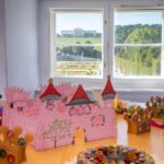 Auf einem Tisch stehen eine rosa Pappburg, verzierte Kronen und Bastelmaterial. Durch das Fenster hat man einen Blick auf einen angelegten Garten und ein großes Gebäude.
