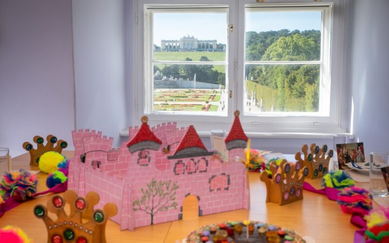 Auf einem Tisch stehen eine rosa Pappburg, verzierte Kronen und Bastelmaterial. Durch das Fenster hat man einen Blick auf einen angelegten Garten und ein großes Gebäude.