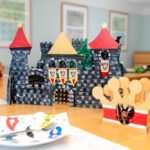 Eine mittelalterliche Spielzeugburg und eine dekorative Krone stehen auf einem mit bunten Servietten gedeckten Holztisch in einem hell erleuchteten Raum.