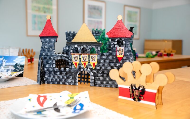 Eine mittelalterliche Spielzeugburg und eine dekorative Krone stehen auf einem mit bunten Servietten gedeckten Holztisch in einem hell erleuchteten Raum.
