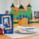 Ein geschmückter Tisch mit einer Papierkrone, bunten Servietten, Silberbesteck und einer Karte mit einer Zeichentrickfigur vor einem grünen Schloss als Tafelaufsatz.