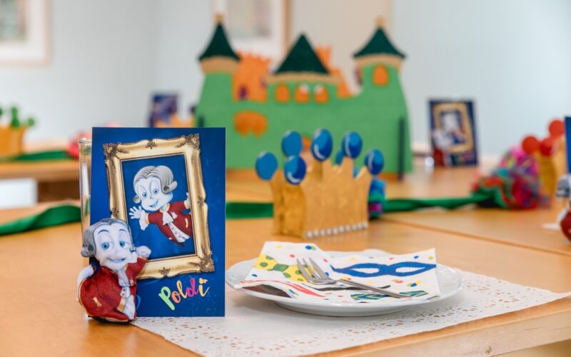 Ein geschmückter Tisch mit einer Papierkrone, bunten Servietten, Silberbesteck und einer Karte mit einer Zeichentrickfigur vor einem grünen Schloss als Tafelaufsatz.