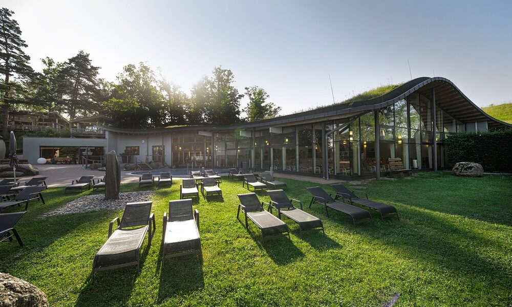 Modernes Gebäude mit großen Glasfenstern und einem geschwungenen Dach, umgeben von Bäumen, mit mehreren leeren Liegestühlen auf einer Wiese davor.