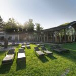 Modernes Gebäude mit großen Glasfenstern und einem geschwungenen Dach, umgeben von Bäumen, mit mehreren leeren Liegestühlen auf einer Wiese davor.