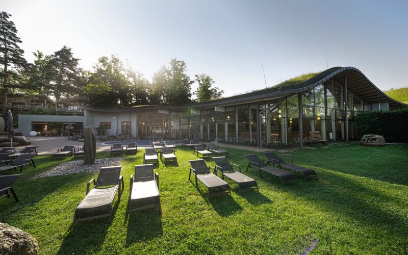Modernes Gebäude mit großen Glasfenstern und einem geschwungenen Dach, umgeben von Bäumen, mit mehreren leeren Liegestühlen auf einer Wiese davor.