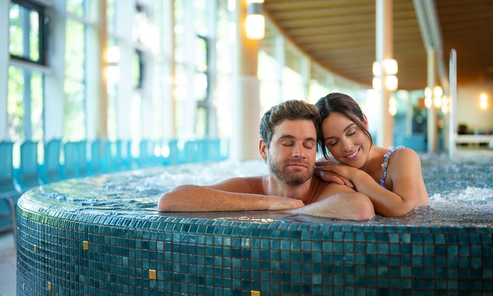 Ein Mann und eine Frau entspannen sich gemeinsam in einem gefliesten Whirlpool, während im Hintergrund natürliches Licht durch große Fenster einfällt.