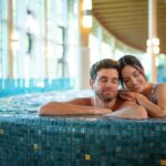 Ein Mann und eine Frau entspannen sich gemeinsam in einem gefliesten Whirlpool, während im Hintergrund natürliches Licht durch große Fenster einfällt.