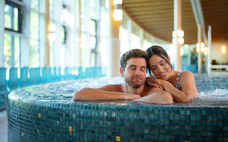 Ein Mann und eine Frau entspannen sich gemeinsam in einem gefliesten Whirlpool, während im Hintergrund natürliches Licht durch große Fenster einfällt.