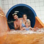 Ein Mann, eine Frau und ein junges Mädchen verlassen gemeinsam eine Indoor-Wasserrutsche, lächeln und plätschern in das Becken am Boden.