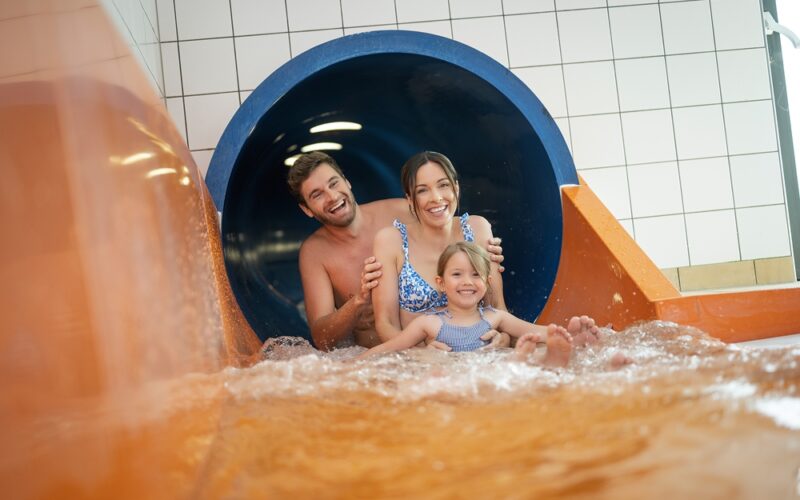 Ein Mann, eine Frau und ein junges Mädchen verlassen gemeinsam eine Indoor-Wasserrutsche, lächeln und plätschern in das Becken am Boden.