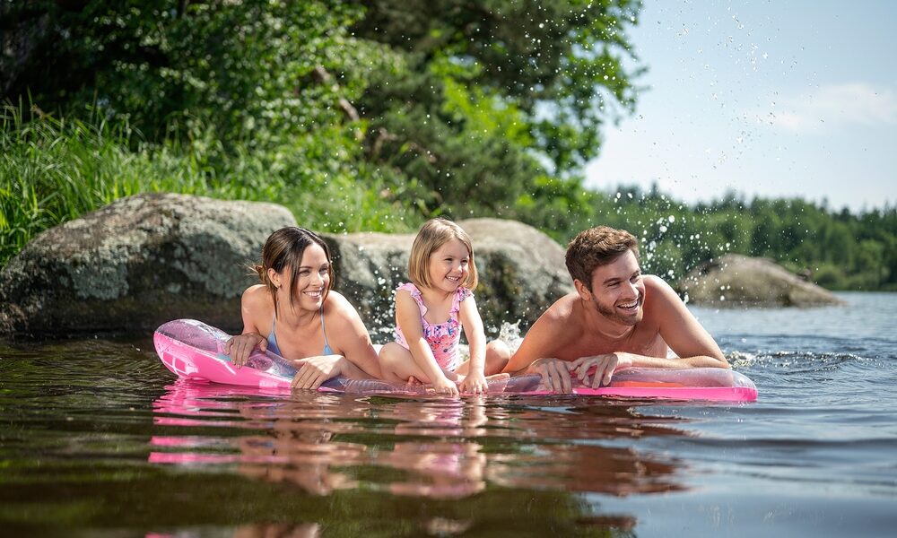 Drei Personen, zwei Erwachsene und ein Kind, liegen auf einem rosafarbenen aufblasbaren Floß in einem See in der Nähe von Felsen und Bäumen, lächeln und spritzen Wasser.