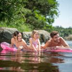 Drei Personen, zwei Erwachsene und ein Kind, liegen auf einem rosafarbenen aufblasbaren Floß in einem See in der Nähe von Felsen und Bäumen, lächeln und spritzen Wasser.