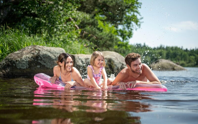 Drei Personen, zwei Erwachsene und ein Kind, liegen auf einem rosafarbenen aufblasbaren Floß in einem See in der Nähe von Felsen und Bäumen, lächeln und spritzen Wasser.