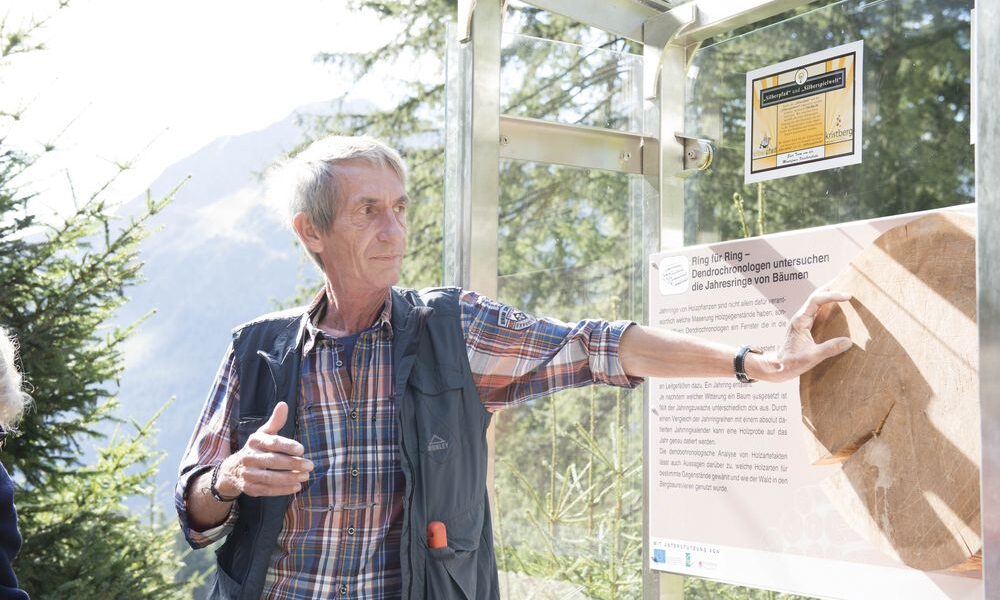 Ein älterer Mann steht im Freien und zeigt auf eine Informationstafel mit Text und einem Fossilienabdruck, mit Bäumen und Bergen im Hintergrund.
