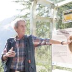 Ein älterer Mann steht im Freien und zeigt auf eine Informationstafel mit Text und einem Fossilienabdruck, mit Bäumen und Bergen im Hintergrund.