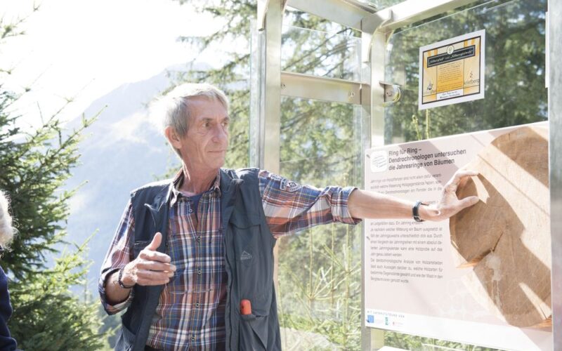 Ein älterer Mann steht im Freien und zeigt auf eine Informationstafel mit Text und einem Fossilienabdruck, mit Bäumen und Bergen im Hintergrund.