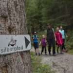 An einem Baum ist ein Schild mit der Aufschrift "Silberpfad" und einem Gnom abgebildet; im Hintergrund geht eine Gruppe von fünf Personen auf einem Waldweg.