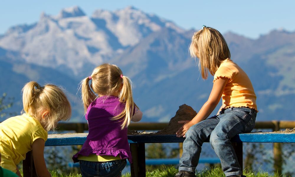 Drei junge Mädchen mit blondem Haar spielen zusammen auf einer Bank im Freien mit Bergen und blauem Himmel im Hintergrund.